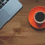 Cup of coffee on an orange saucer next to a laptop on a wooden table.