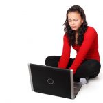 A woman in a red sweater is writing a blog using a laptop on the floor.