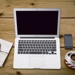 A laptop, accompanied by a notebook with a pen, a smartphone, and a cup of coffee on a saucer.